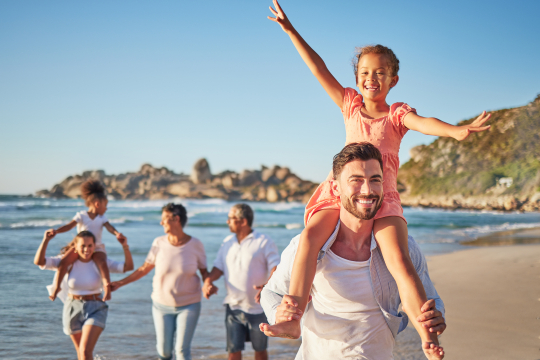 Vorne ein Vater mit einem Mädchen auf den Schultern, links daneben eine Mutter mit einem weiteren Kind, und Oma und Opa am Strand laufend